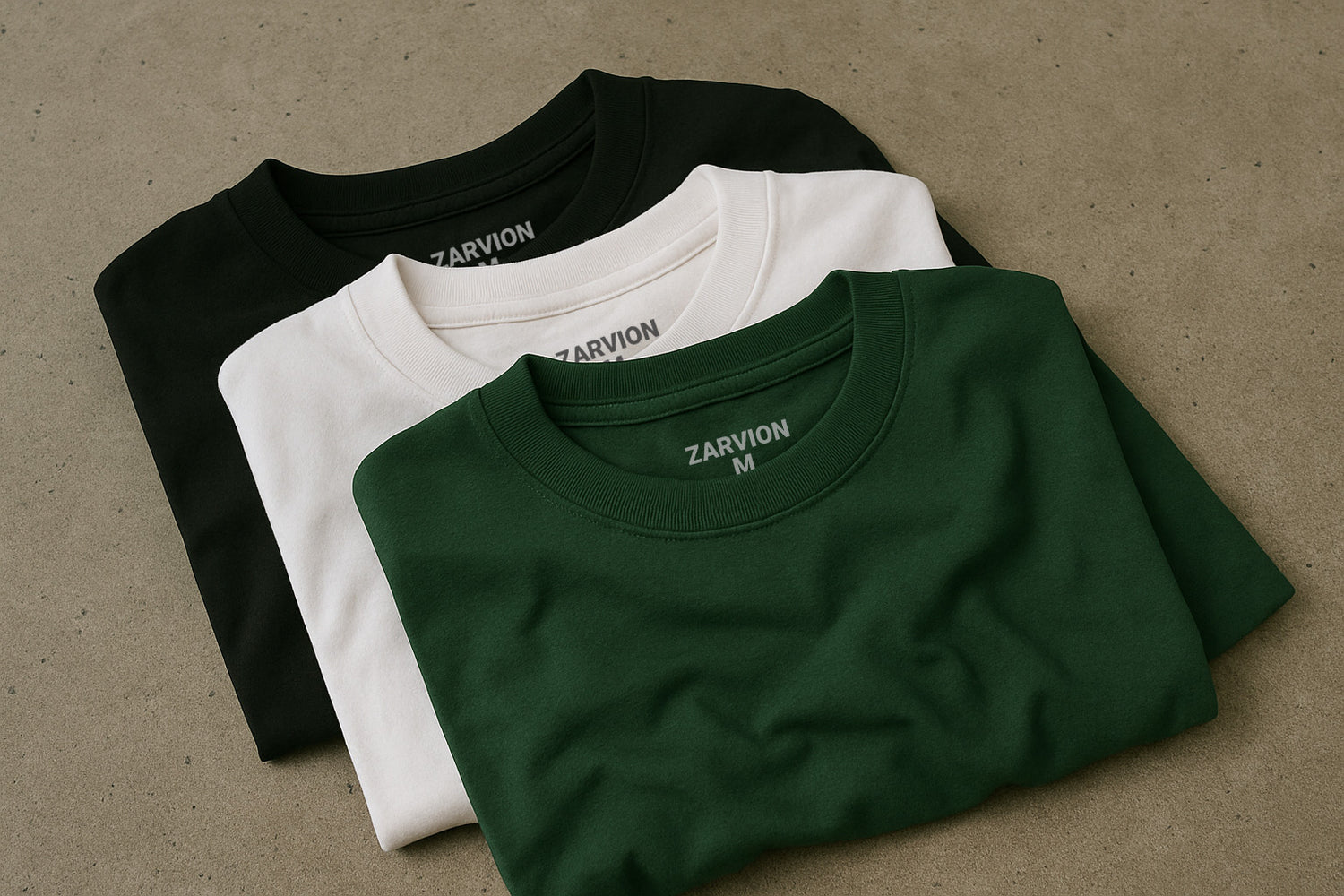 Three folded crewneck t-shirts in green, white, and black stacked on a concrete surface with light fabric wrinkles – minimalist flat lay style for Zarvion streetwear packs